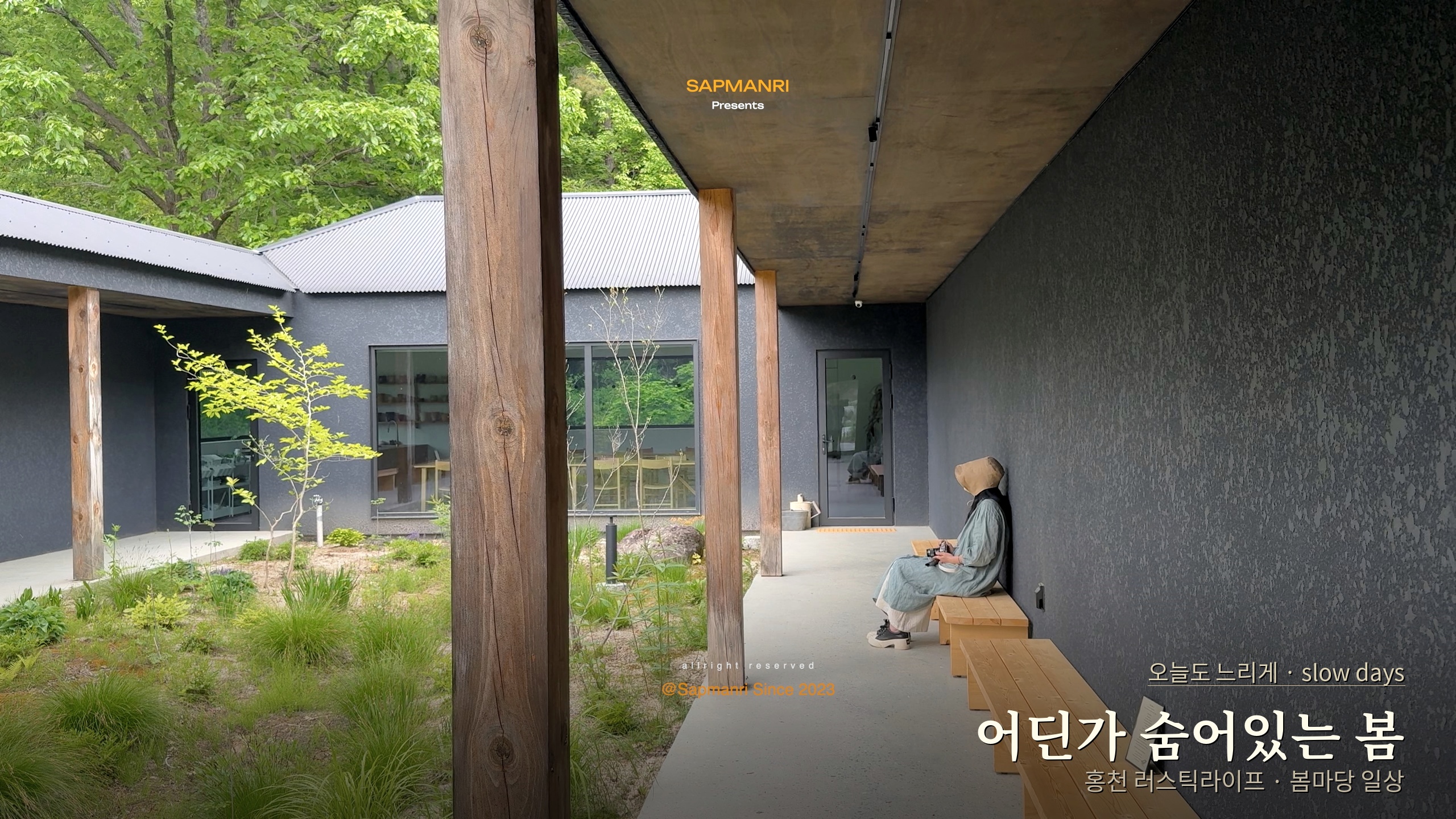 A woman seated on a wooden bench in a dark-walled courtyard filled with fresh green plants.