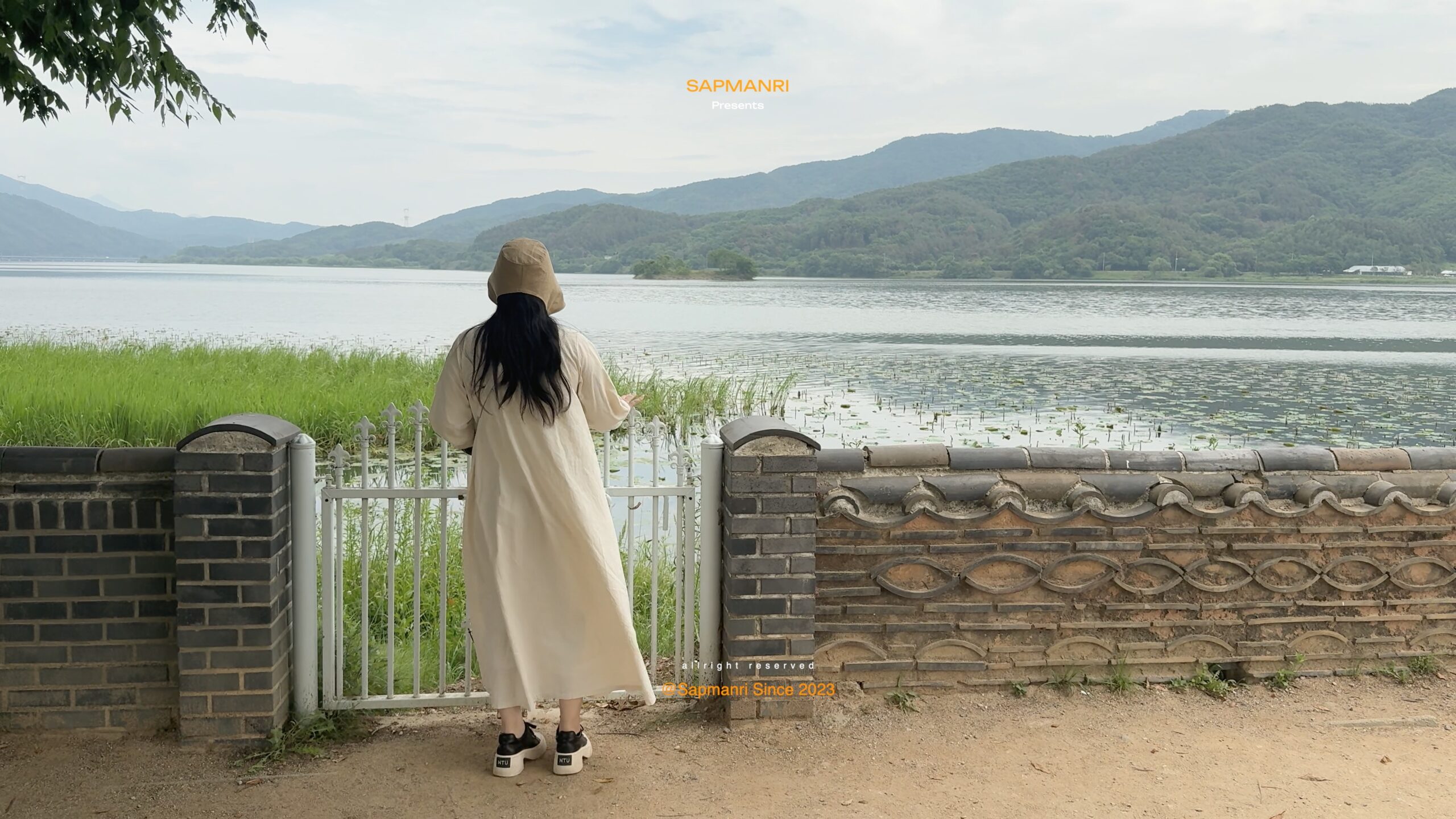 A person in a cream coat standing beside calm water and green mountains at Dumulmeori.