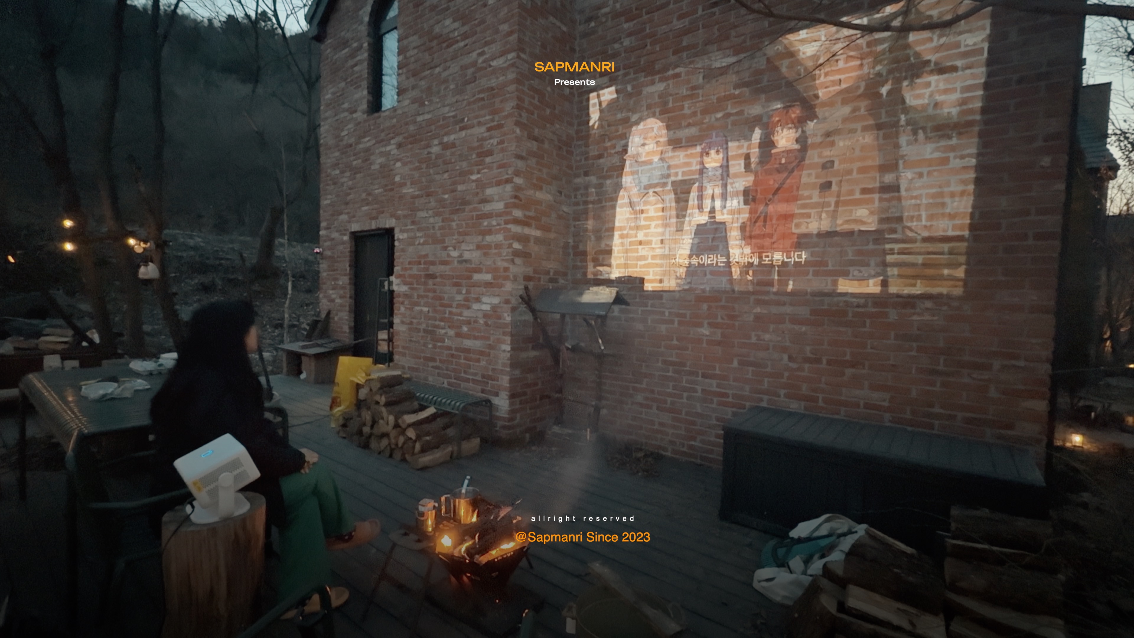 A video projected onto a brick wall while a person sits near a small open brazier on the wooden deck.
