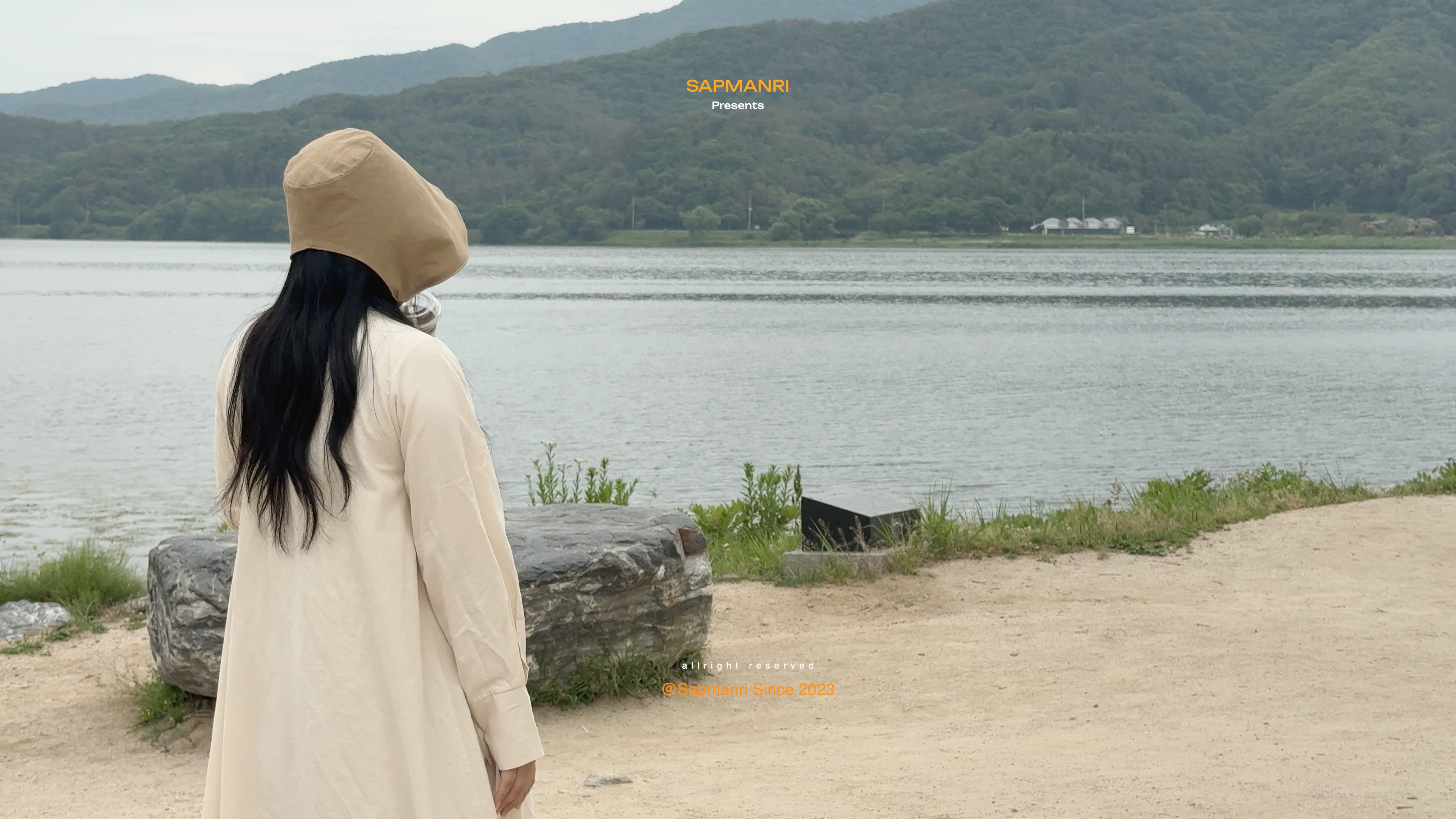 A person in a cream coat pausing beside the river with wooded mountains across the water.