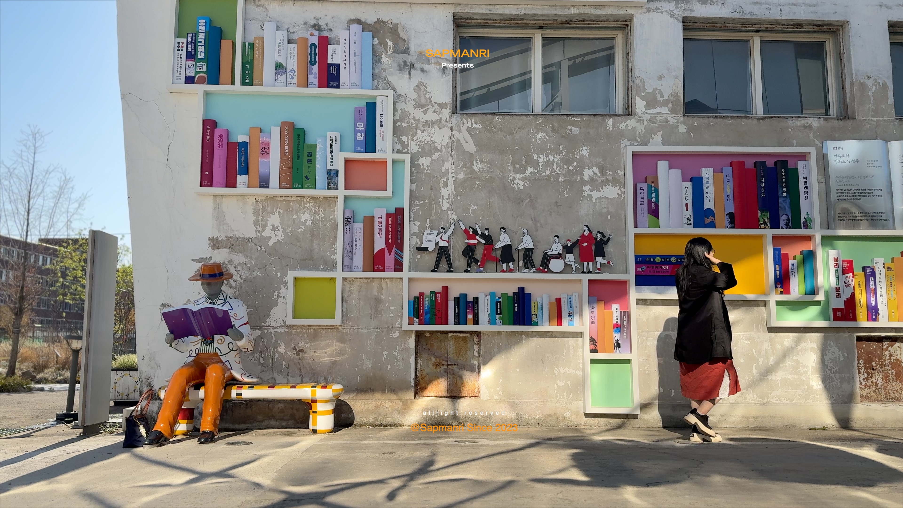 A colorful outdoor book installation on a weathered warehouse wall beside a seated sculpture.