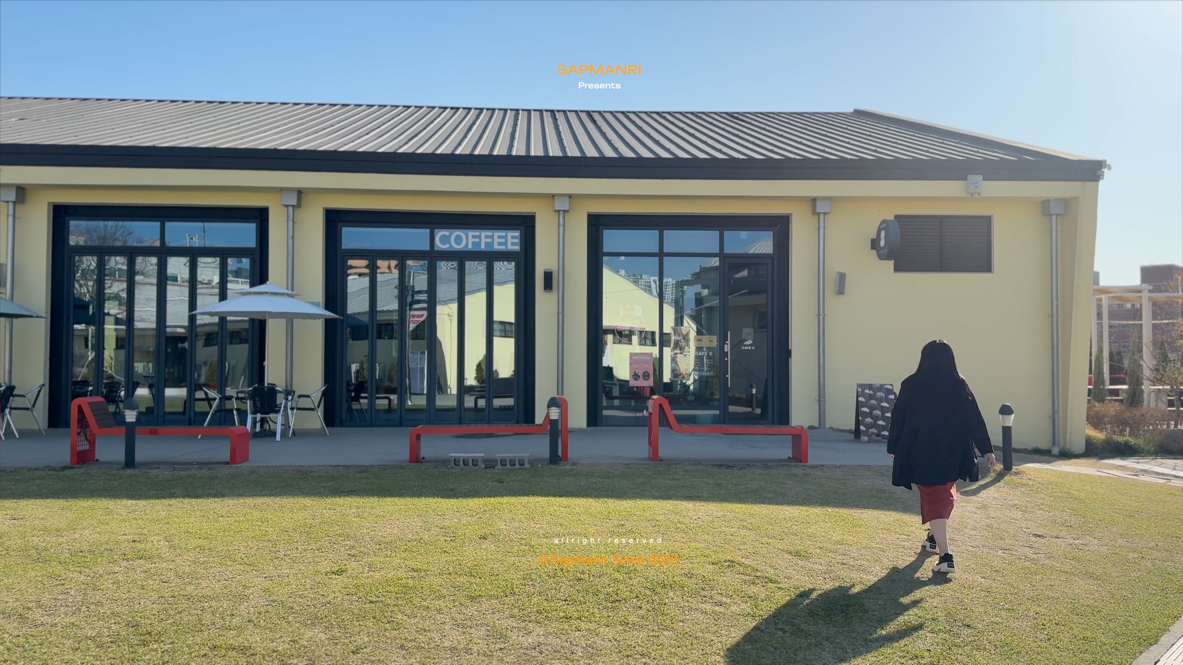 A yellow warehouse building marked Coffee and Cafe C, with red benches and a person crossing the grass.