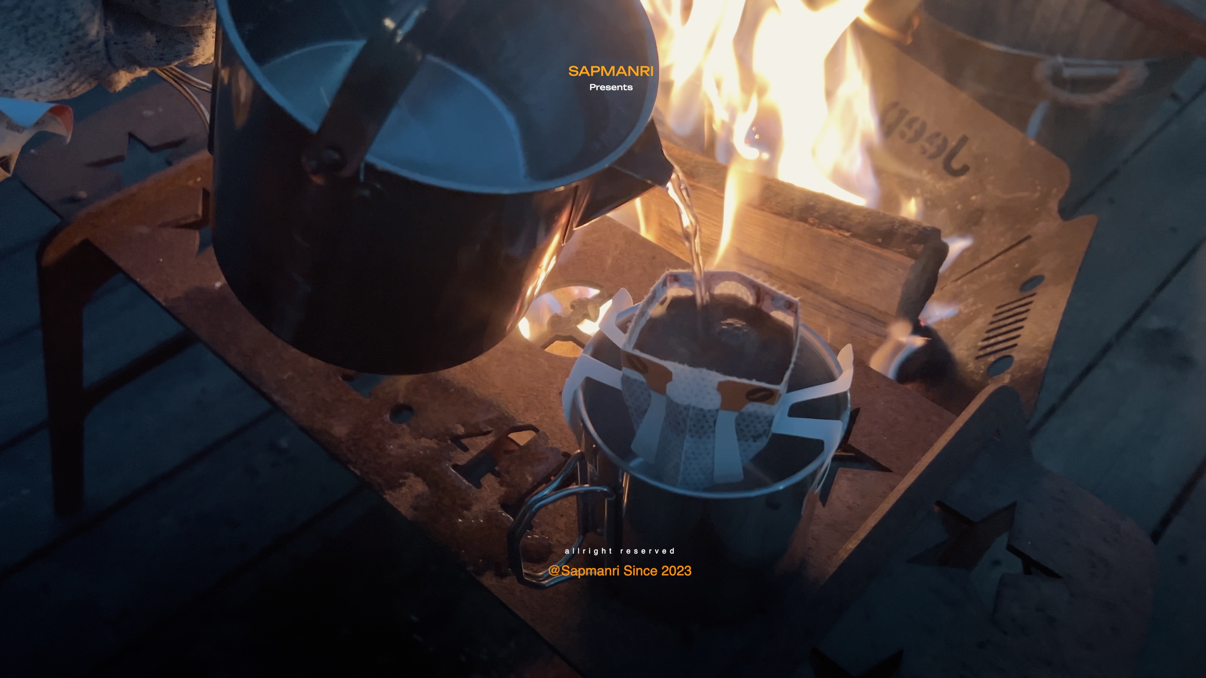 Pour-over coffee equipment set on a low wooden surface close to flames rising from an outdoor brazier.