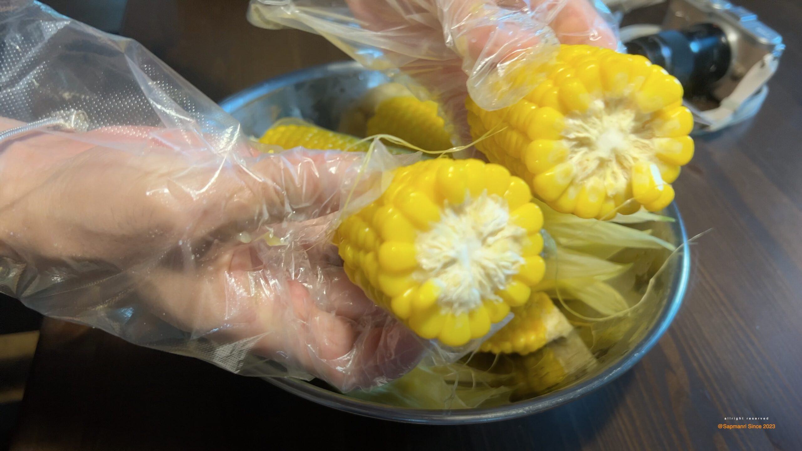 Gloved hands placing cooked corn pieces into a storage bag over a metal bowl.