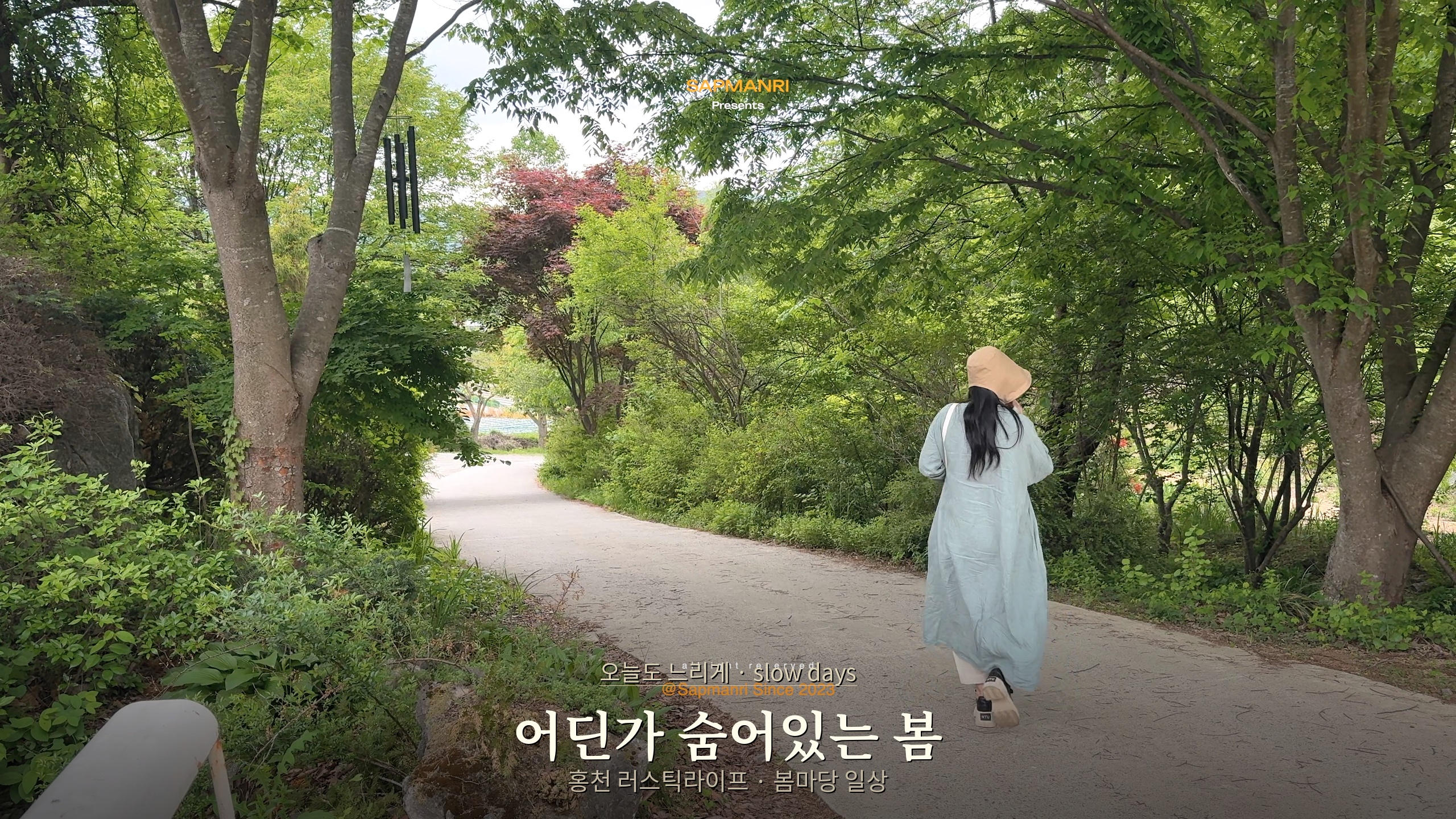 A woman walking away along a narrow paved road under a dense canopy of green trees.