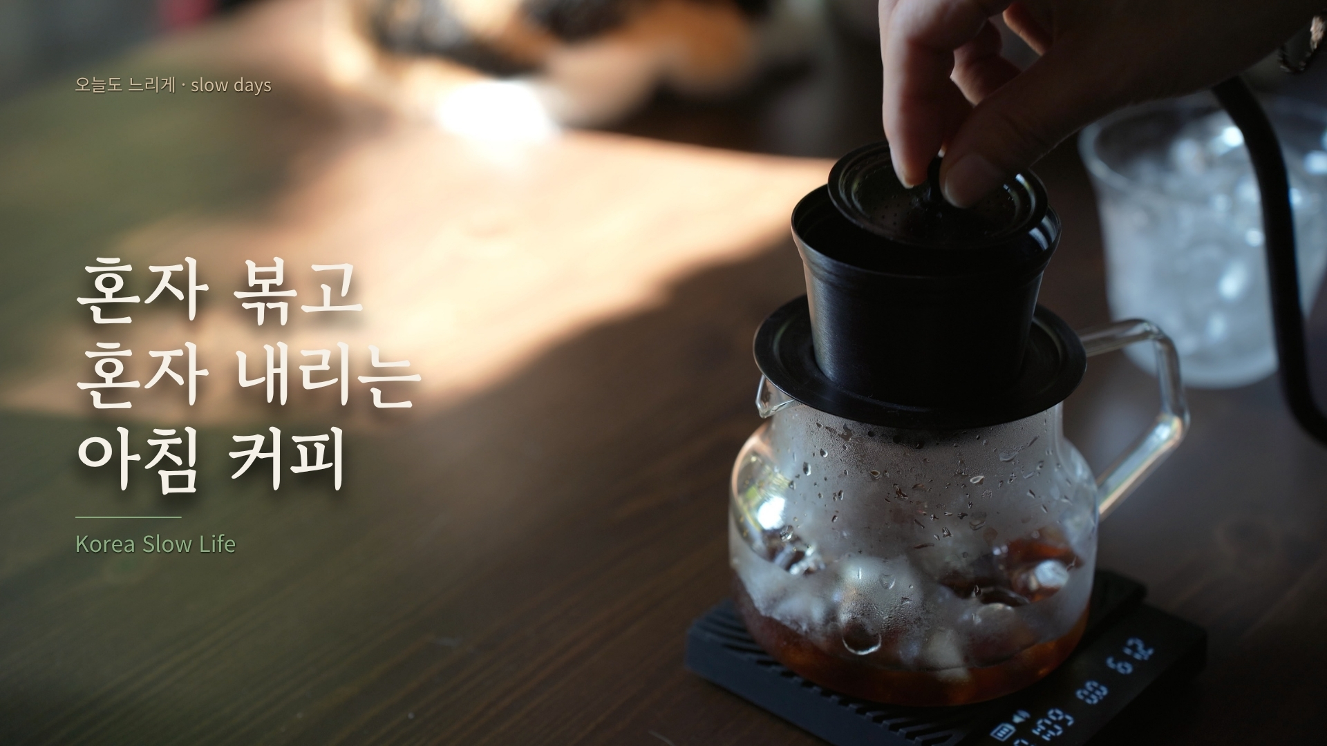 A hand grinding coffee above a glass server and digital scale in morning light.