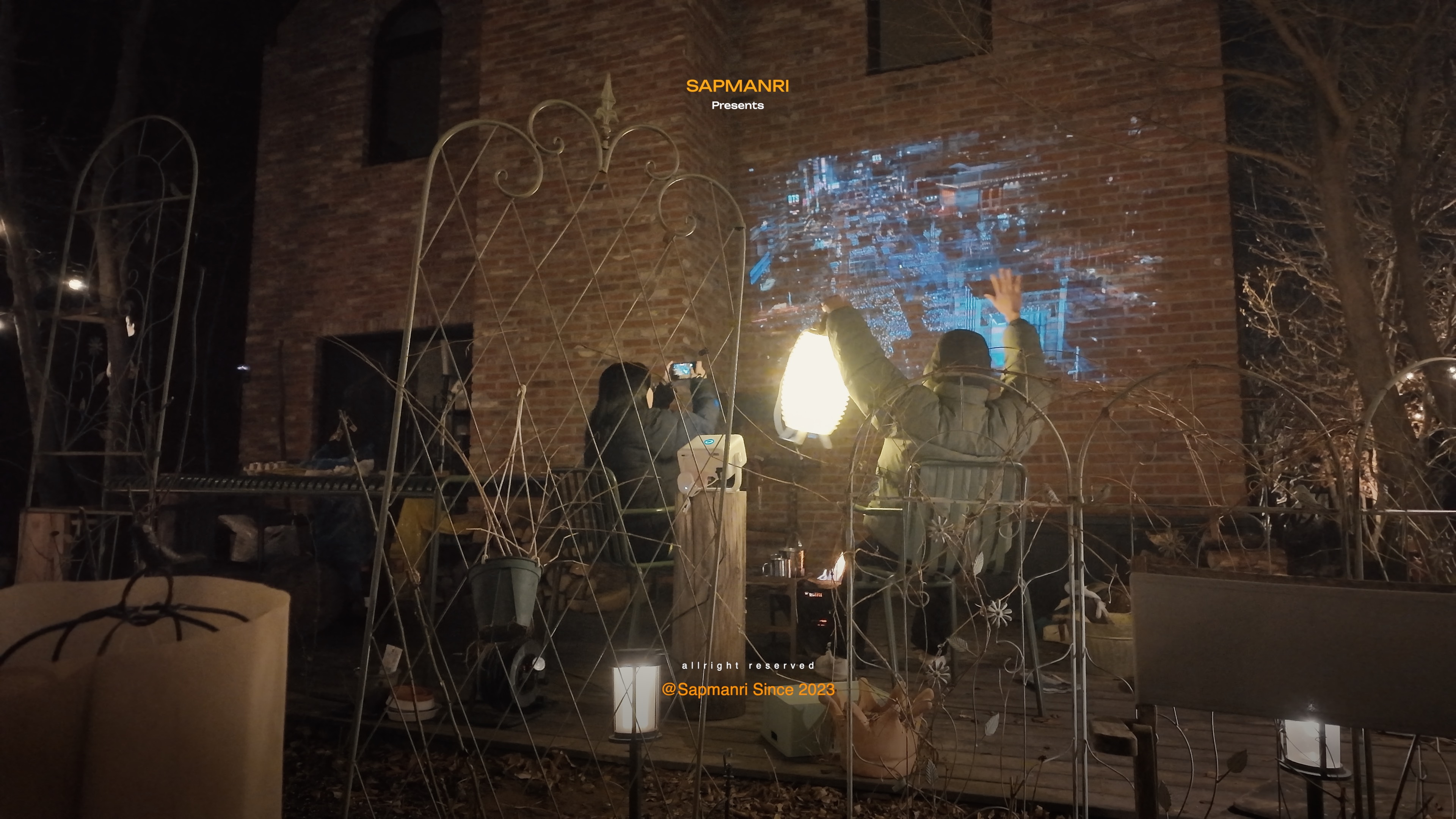 Several people standing and sitting in a dark garden while one person raises both arms toward a wall projection.