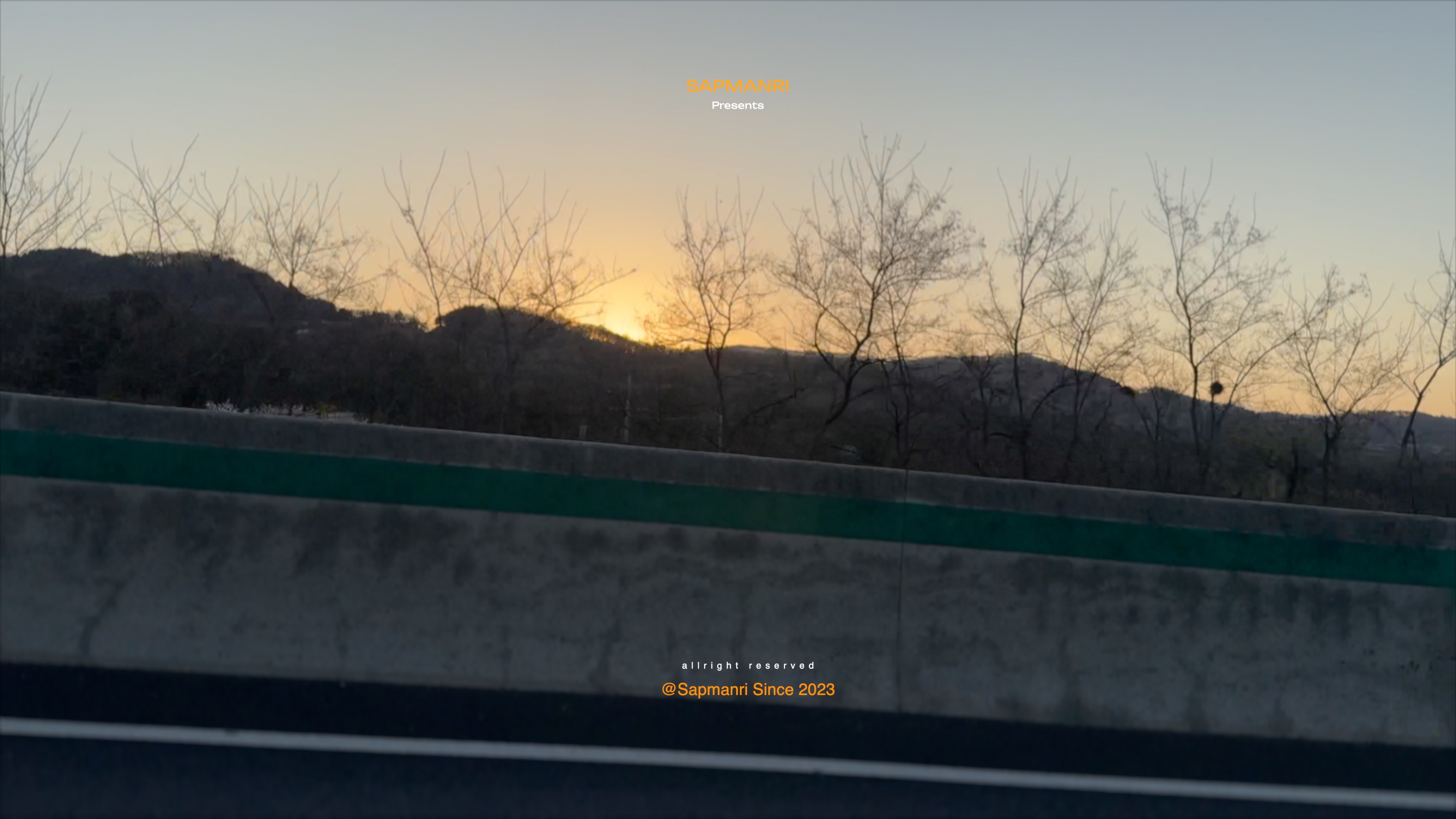 A winter-bare hillside and orange sunset seen past a highway barrier from a moving vehicle.