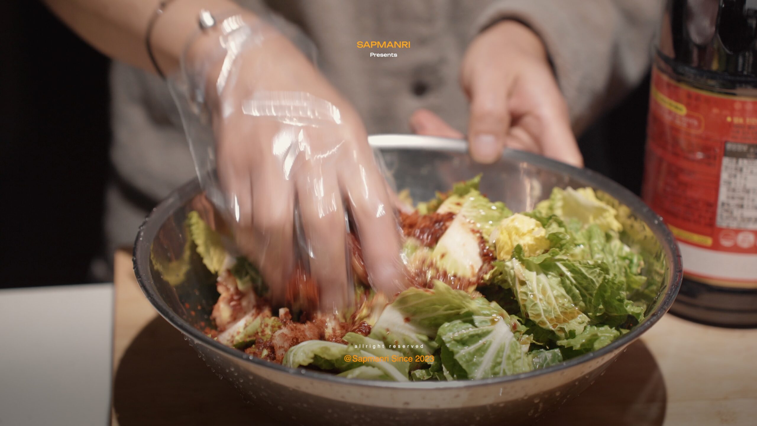 Two hands mixing chopped spring cabbage with red seasoning in a large stainless-steel bowl.