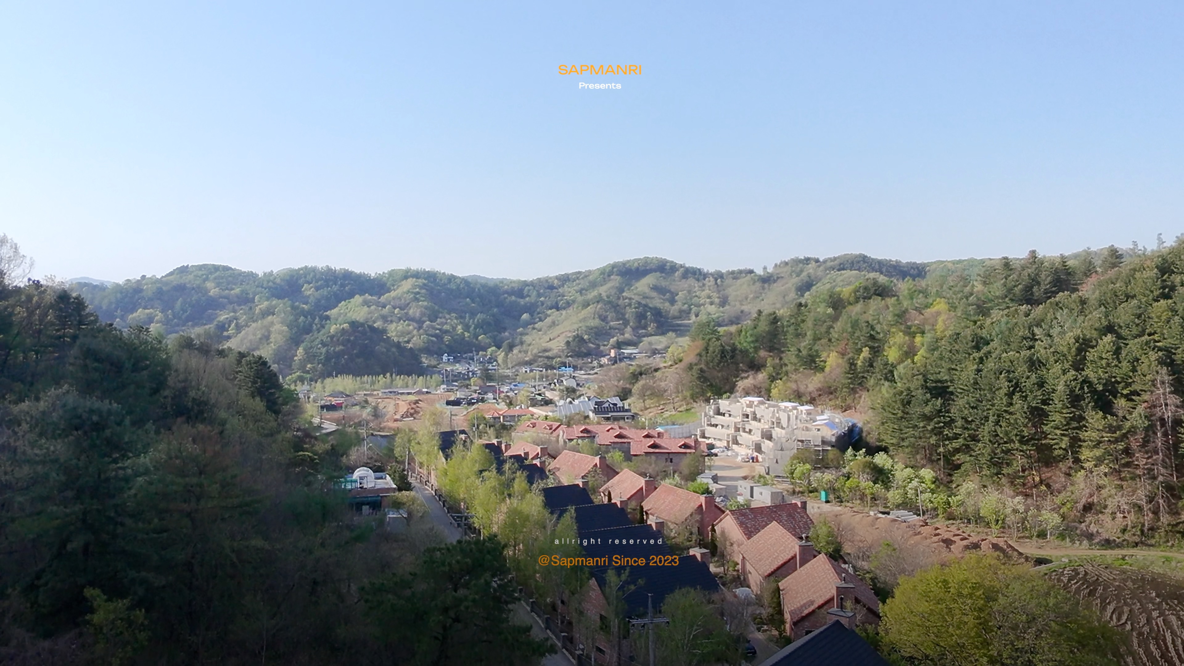 A high view across village roofs toward a forested ridge under soft spring light.