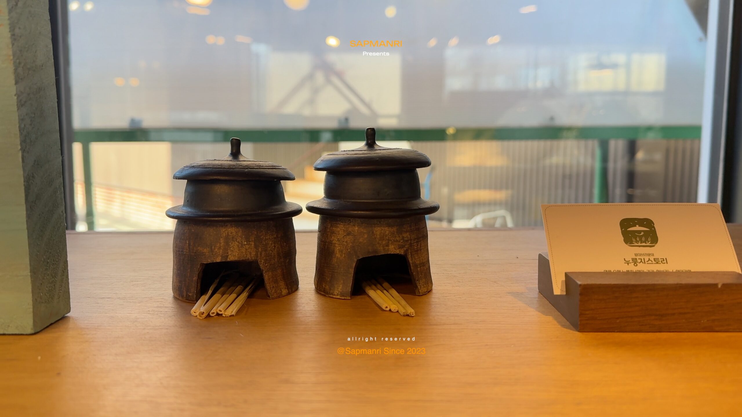 Two small lidded earthen containers with wooden sticks on a restaurant counter by a window.