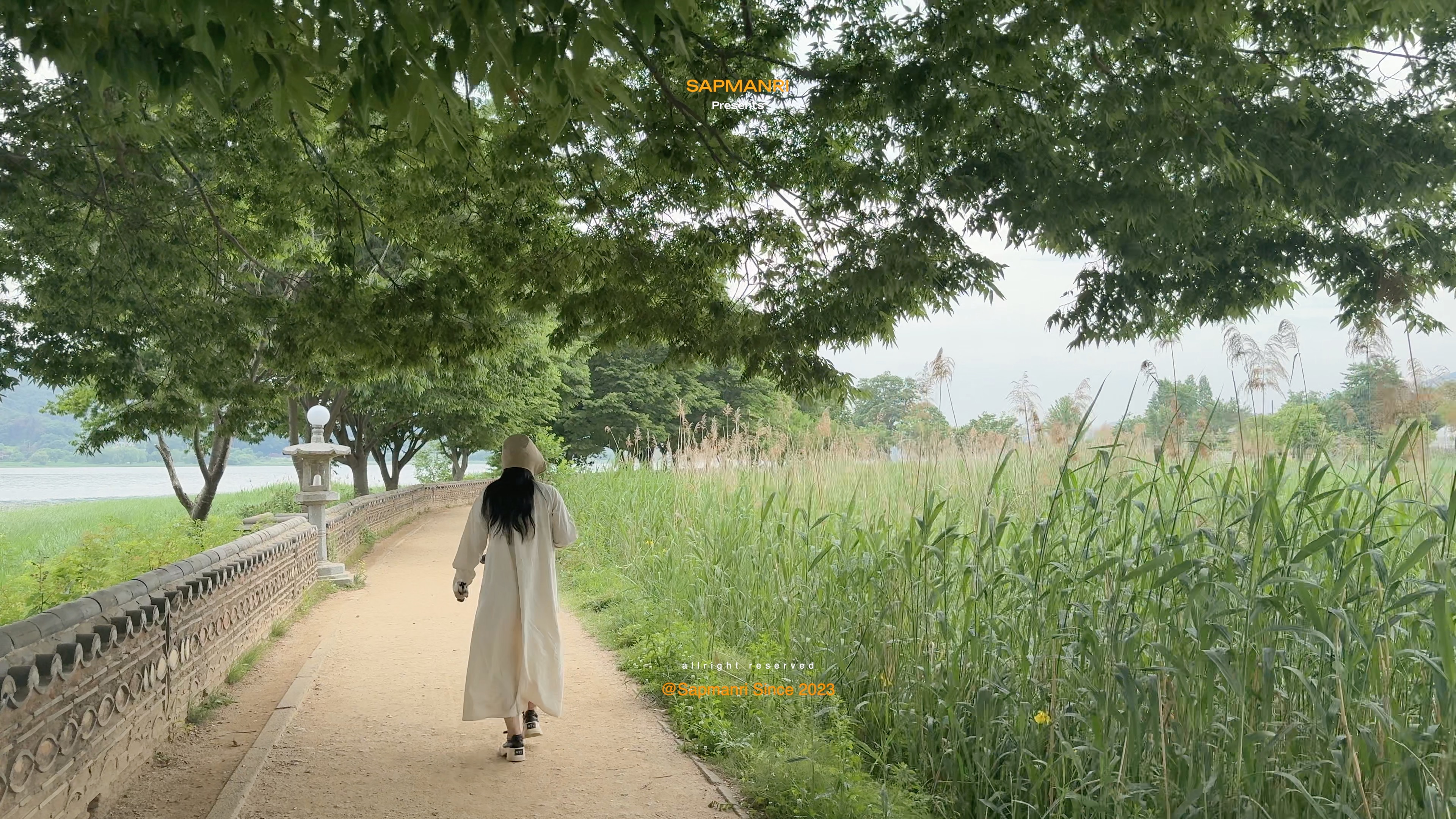 A person in a cream coat walking along an earthen path between a low wall and tall reeds at Dumulmeori.