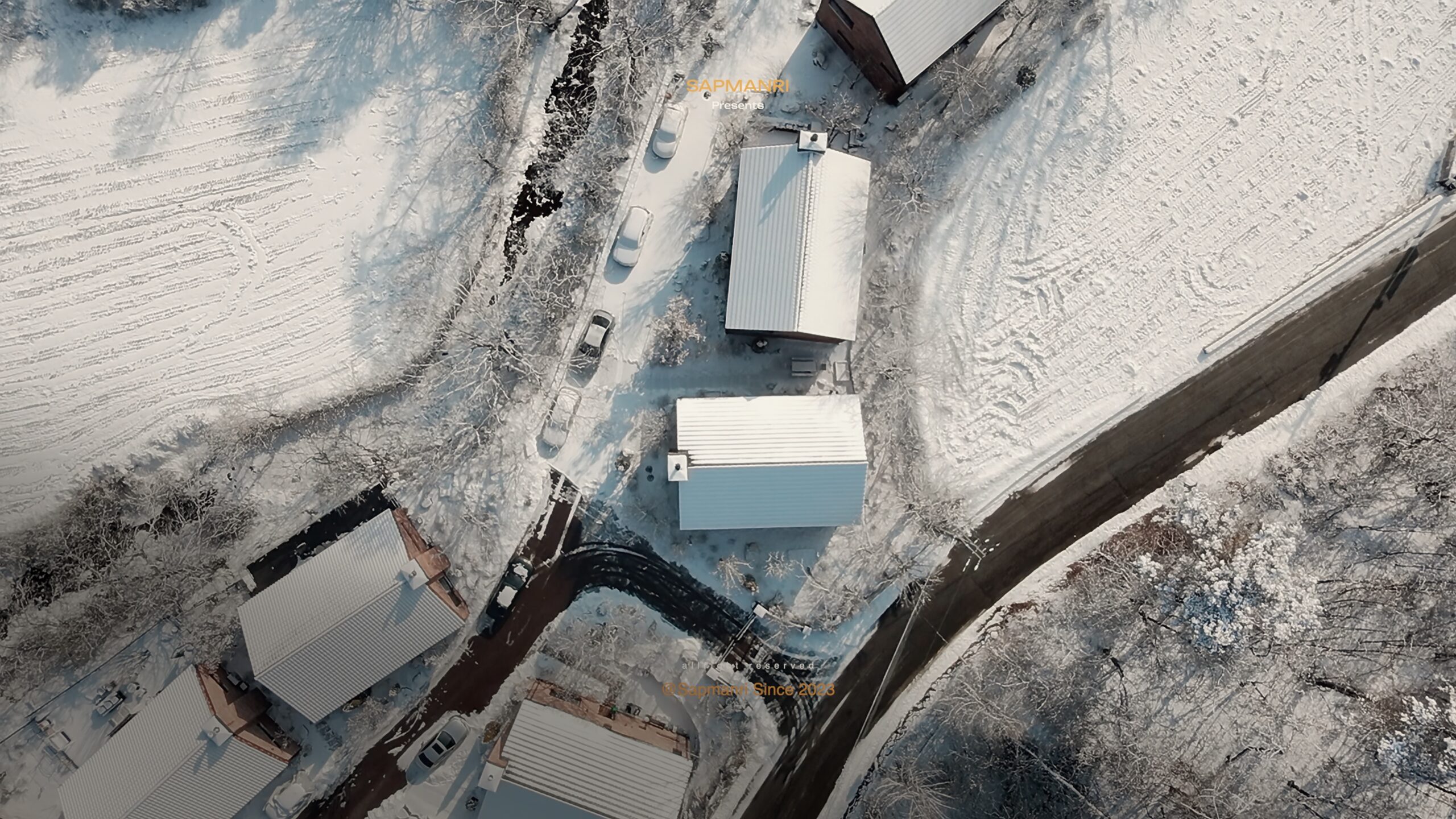 An aerial view of snow-covered village roofs, narrow roads, fields, and dark winter trees.