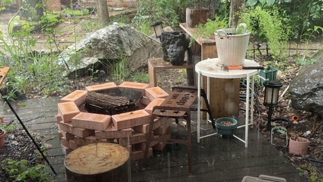 A rain-wet garden patio with a circular brick fire pit, stools, and potted plants.