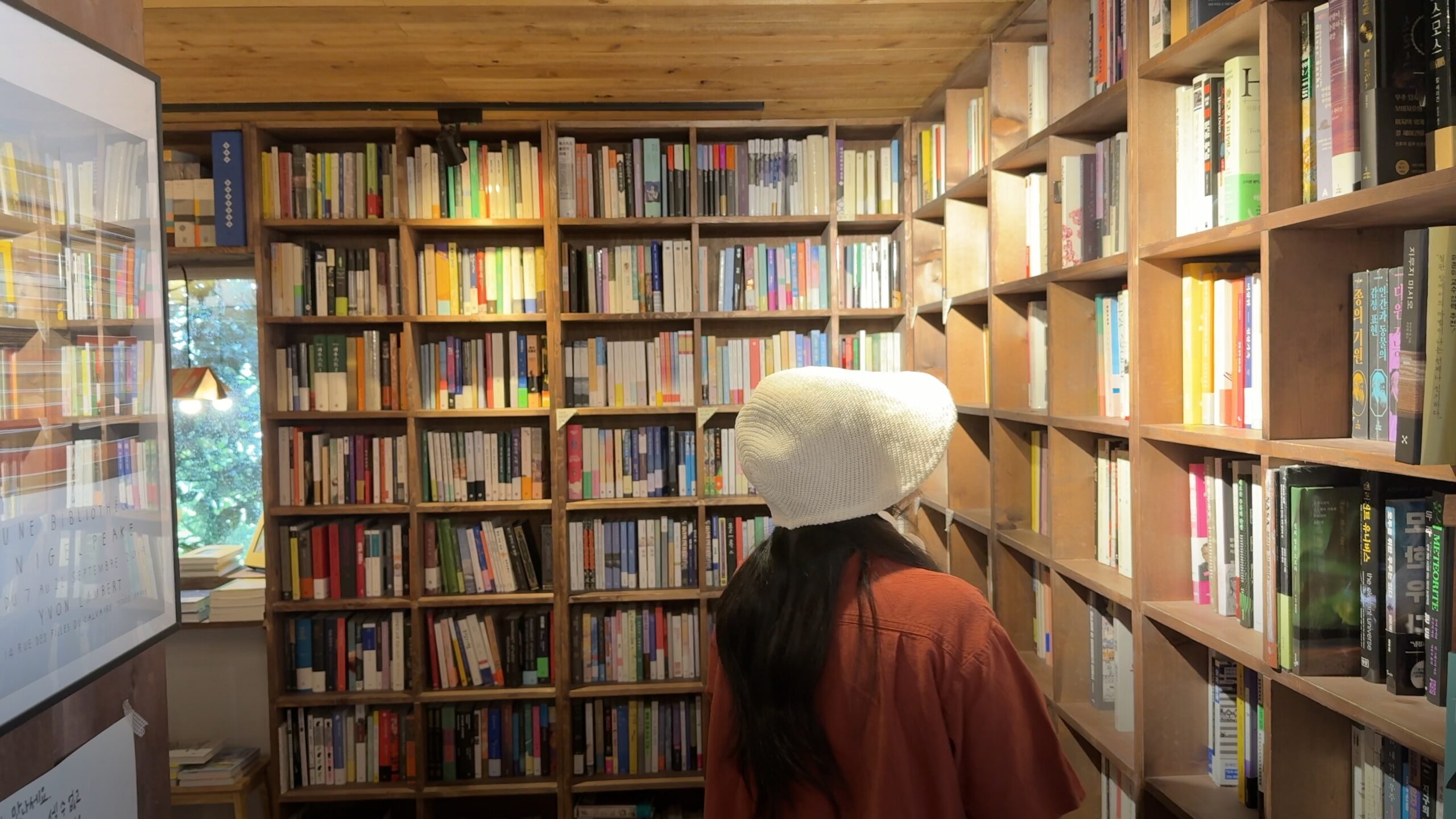 A person in a white bucket hat browsing tall wooden bookshelves in a bookshop.