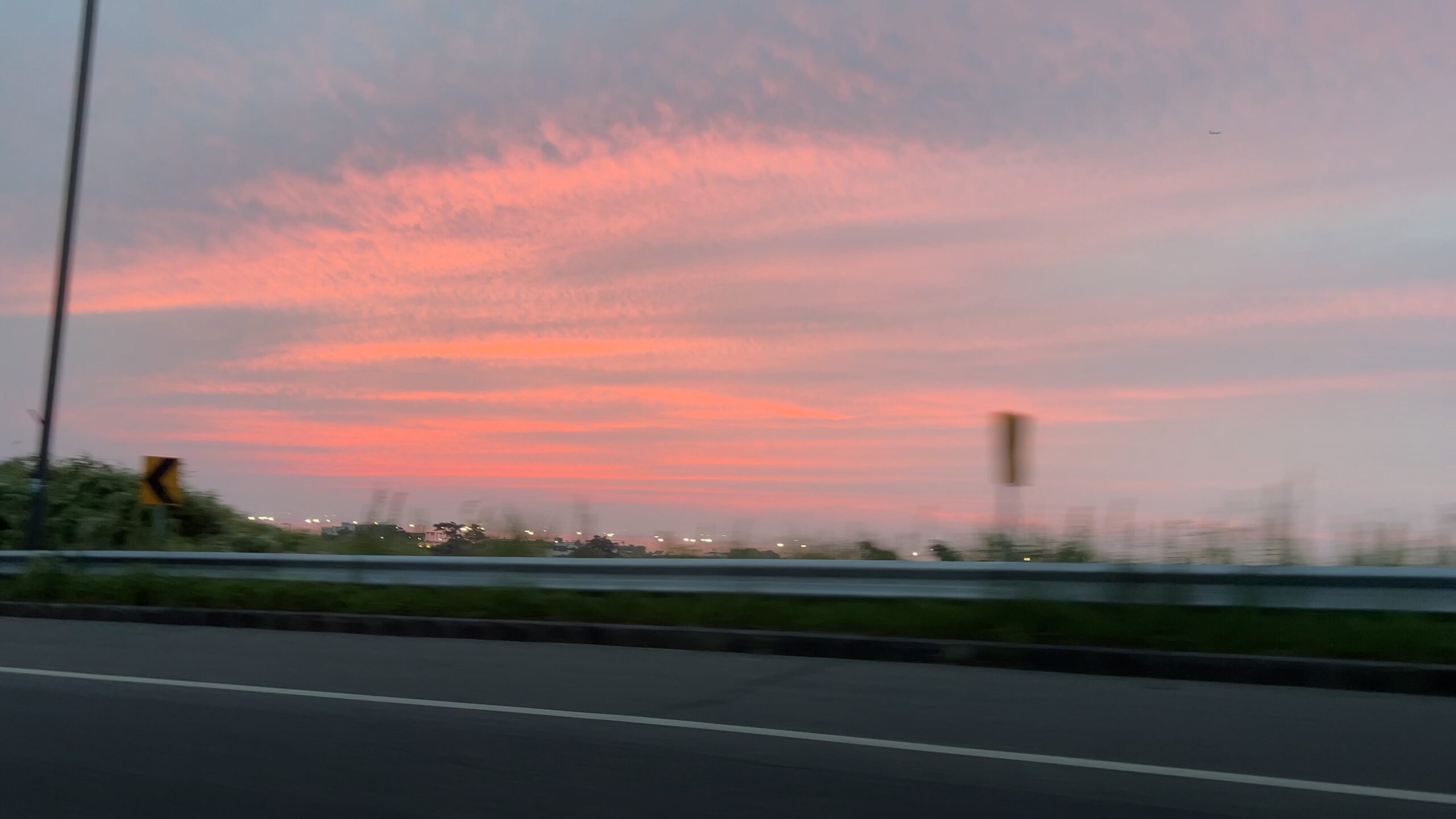 A red and pink sunset beyond a highway, photographed from a moving car.