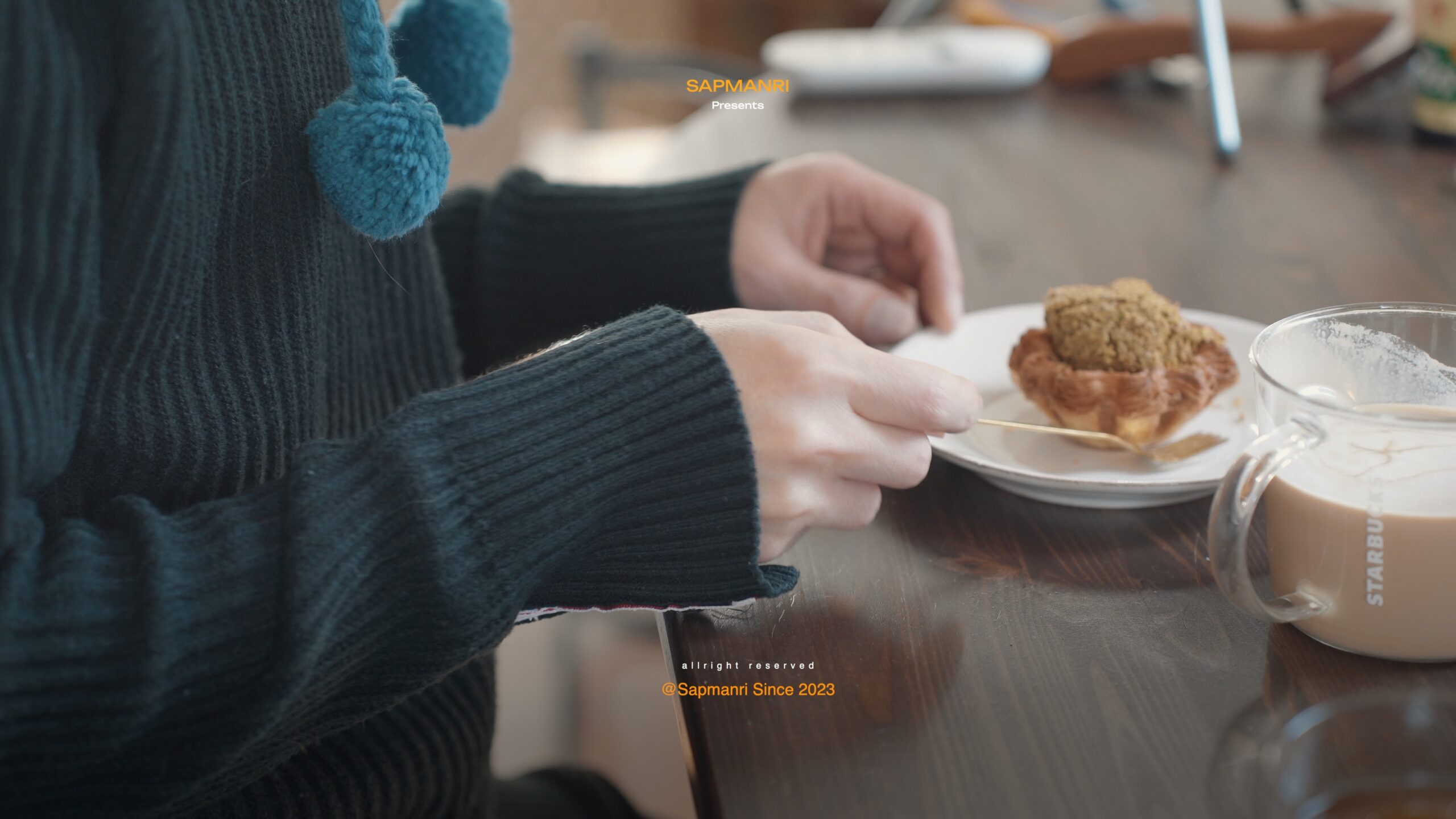 Hands beside a milky coffee and a pastry topped with a small green cake at a wooden table.
