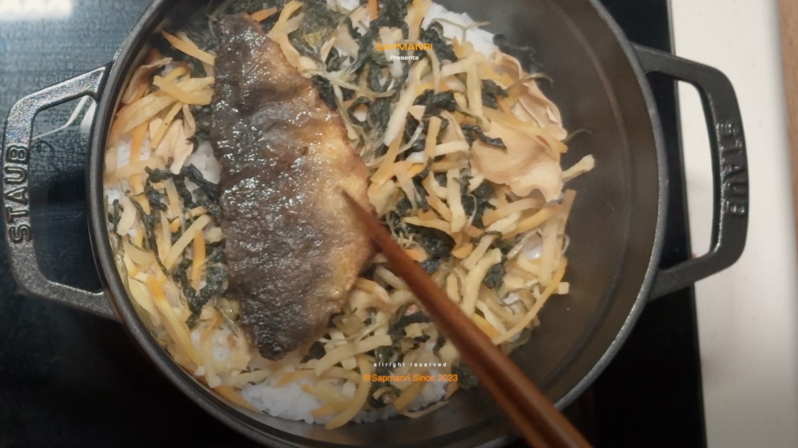 A cast-iron pot holding rice, gondre greens, sliced vegetables, and a large piece of cooked fish.