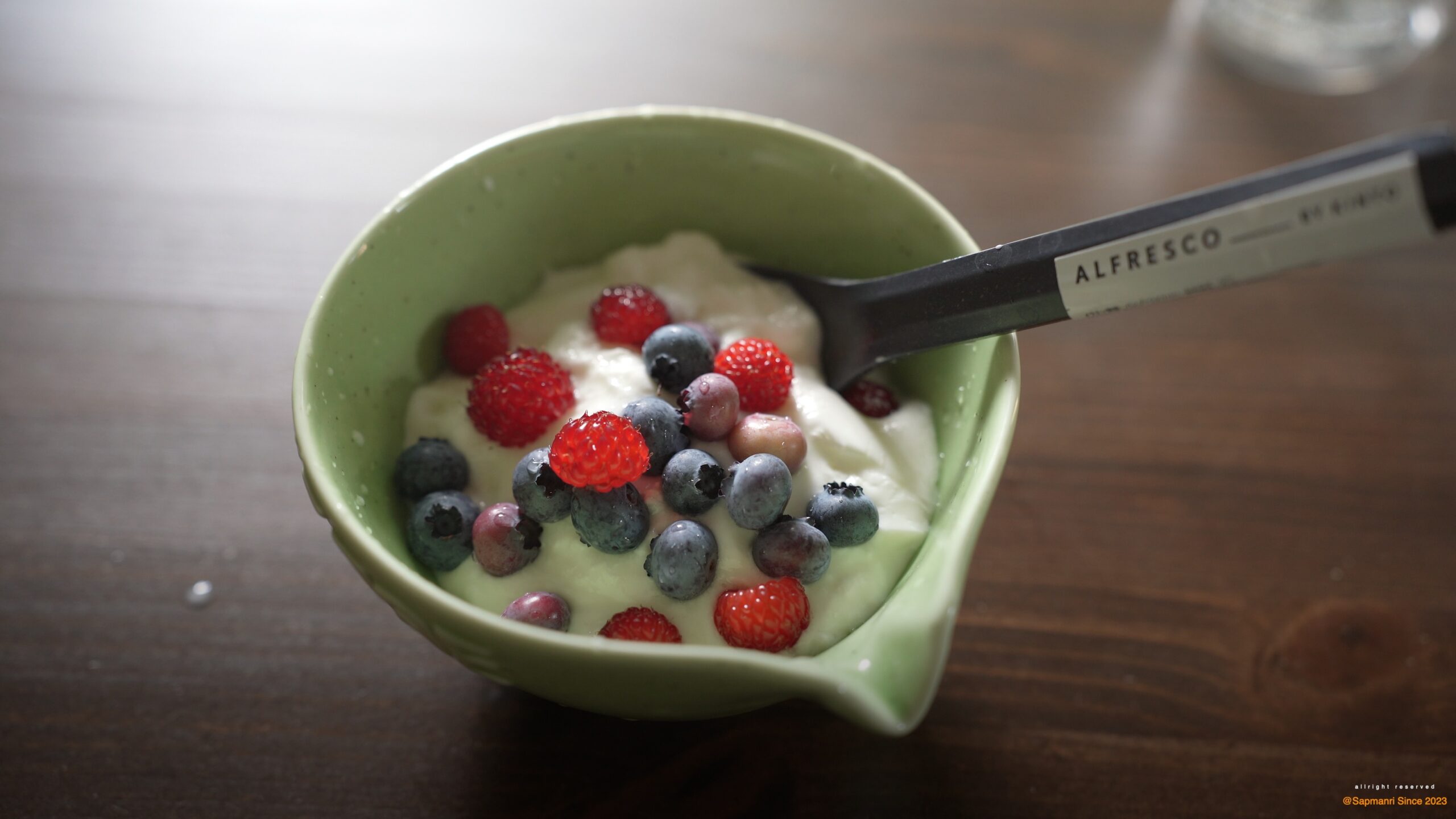 A green bowl of yogurt topped with blueberries and small red berries beside a spoon.