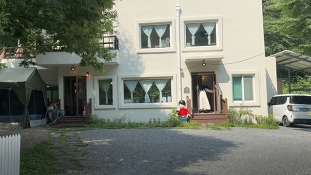 A white two-storey countryside house among trees, with a person standing in one doorway.