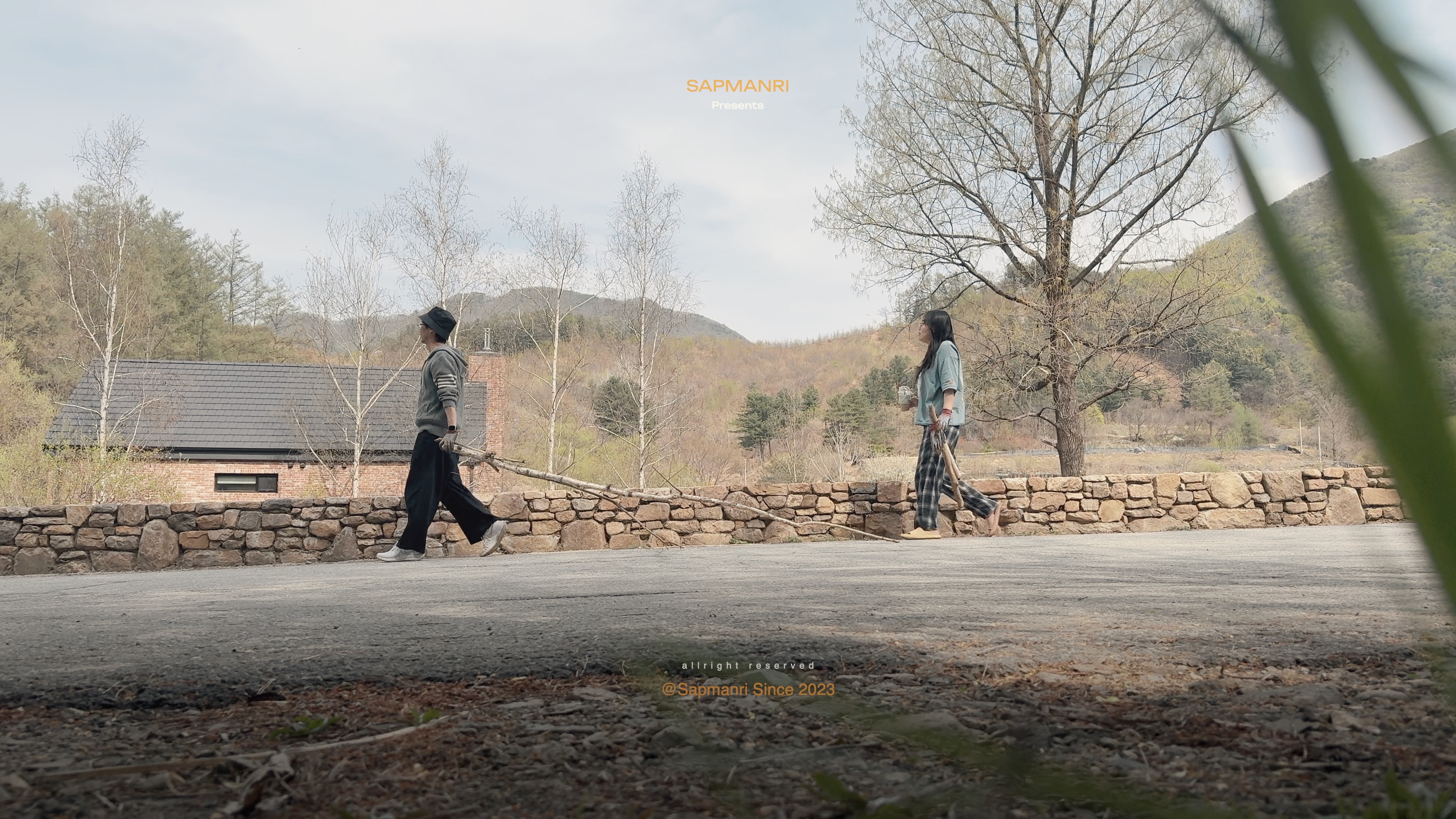 Two people dragging long bare branches along a quiet paved road beside a stone wall.
